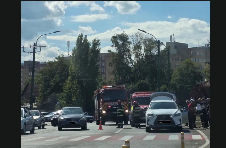O intersecție aglomerată cu mașini, un camion de pompieri, și mai mulți oameni, sugerând un accident rutier. Cerul este parțial înnorat.