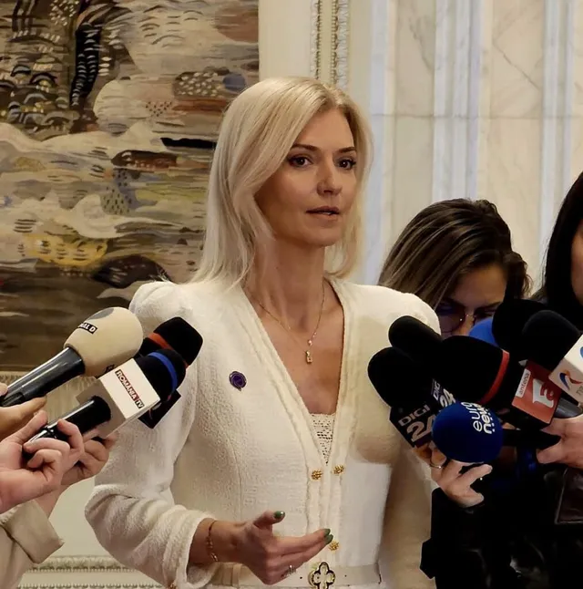 O femeie cu părul blond, purtând o bluză albă, stă în fața mai multor microfoane de presă.