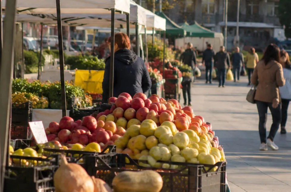 Diverse fructe sunt expuse în coșuri de plastic sub un prețar, în timp ce oameni merg pe o esplanadă însorită în fundal.