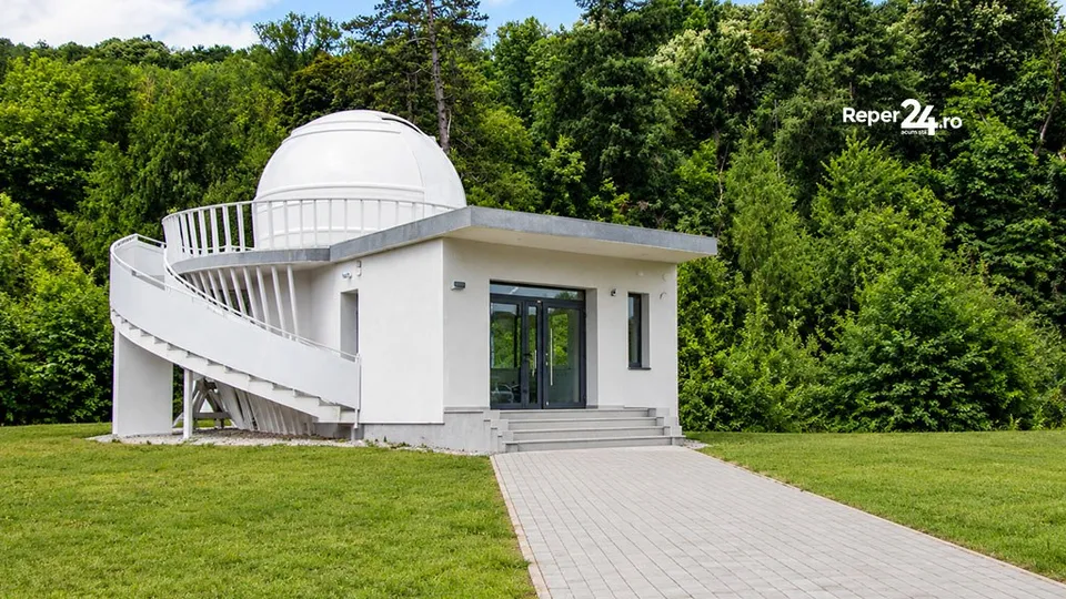 O clădire albă cu o cupolă rotundă și o scară spirală exterioară este situată pe un teren cu iarbă lângă o pădure densă.