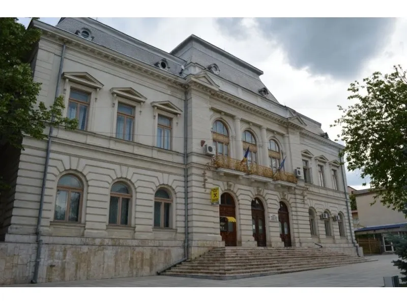 O clădire cu fațadă de culoare deschisă prezintă ferestre înalte, un balcon cu balustradă și scări largi la intrare sub un cer înnorat.
