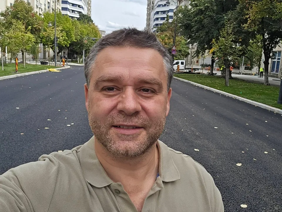 Un bărbat cu părul scurt și barbă stufoasă poartă un tricou polo de culoare bej în fața unui drum asfaltat și a unor copaci verzi.