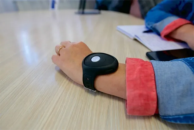 O persoană poartă un dispozitiv electronic negru pe încheietura mâinii, stând cu brațul întins pe o masă de lemn lângă un caiet și un telefon.