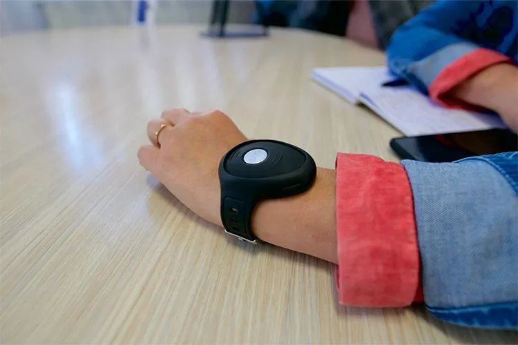 O persoană poartă un dispozitiv electronic negru pe încheietura mâinii, stând cu brațul întins pe o masă de lemn lângă un caiet și un telefon.