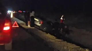 Un autoturism gri este poziționat într-un șanț lângă o stradă, în timp ce persoane și vehicule cu lumini aprinse sunt vizibile în fundal noaptea.