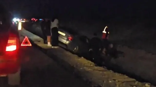 Un autoturism gri este poziționat într-un șanț lângă o stradă, în timp ce persoane și vehicule cu lumini aprinse sunt vizibile în fundal noaptea.