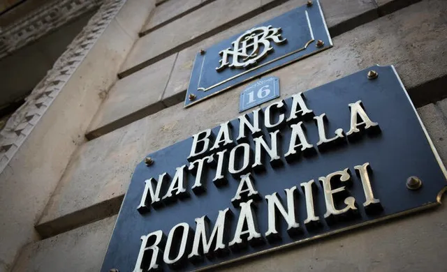Un panou negru cu textul BANCA NATIONALA A ROMANIEI este montat pe o fațadă de piatră lângă un logo BNR.