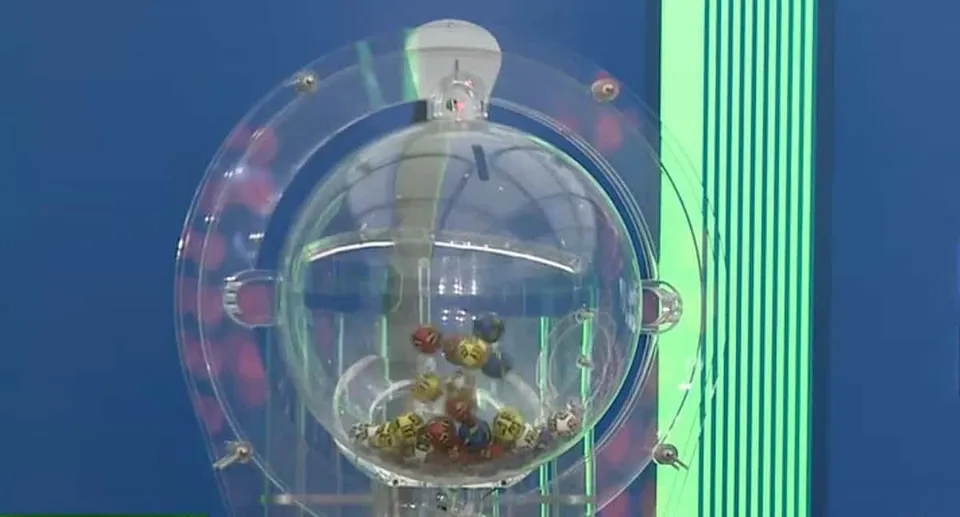 Un aparat de tragere transparent conține numeroase bile colorate în interiorul unei sfere de sticlă montate pe un suport vertical.