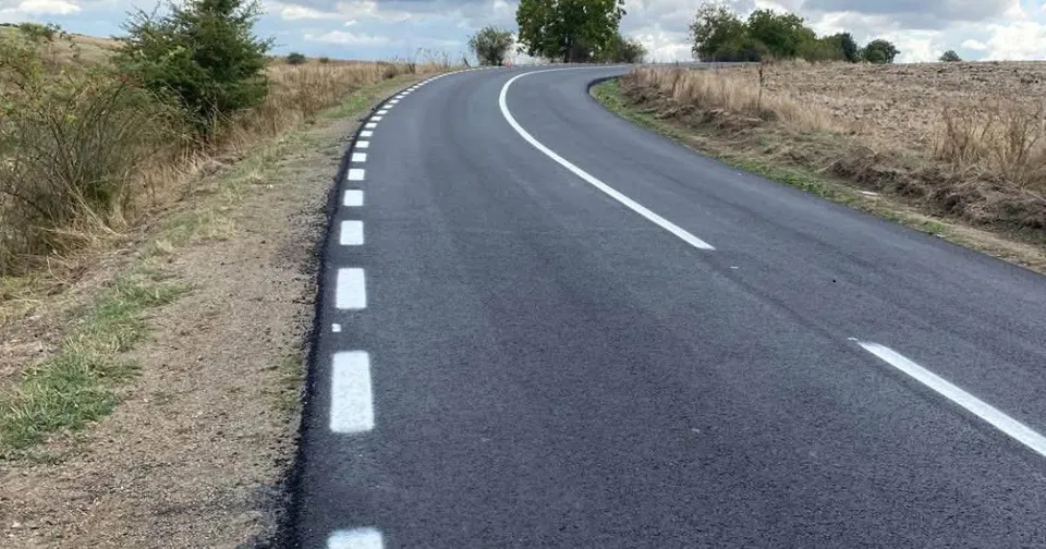 O porțiune de drum asfaltat cu marcaje rutiere albe curbează spre dreapta printre câmpuri și vegetație sub un cer înnorat.