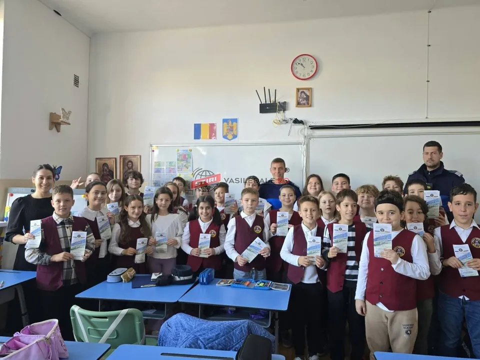 Un grup de elevi stă în fața unor mese de clasă, ținând broșuri albe, alături de câțiva adulți într-o încăpere cu ceas pe perete.