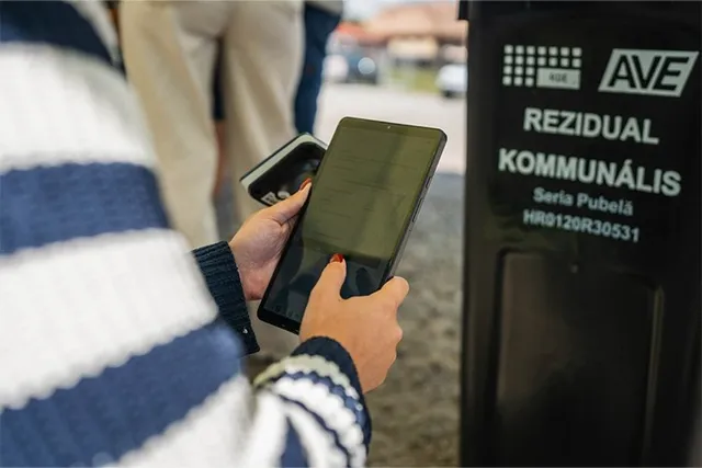 O persoană ține un dispozitiv electronic deasupra unui recipient de deșeuri negru pe care este scris text alb.