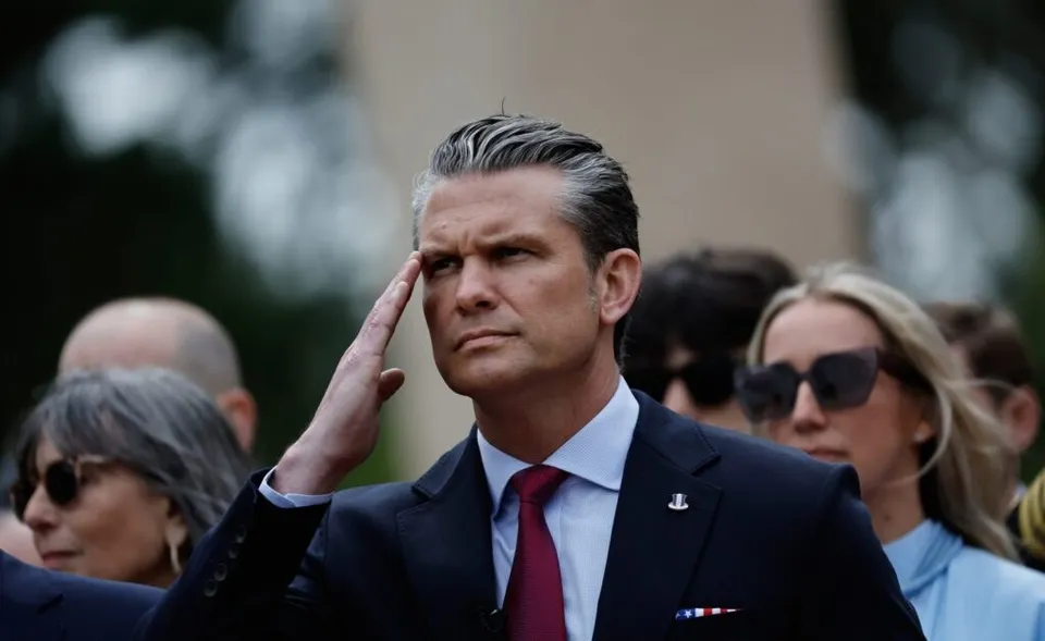 A man in a dark suit and red tie salutes outdoors, surrounded by people in formal attire.