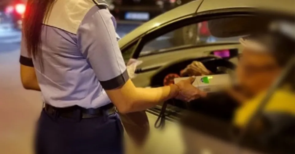 O polițistă în uniformă ține un aparat de testare alcoolemiei în interiorul unui autoturism spre un bărbat aflat la volan.