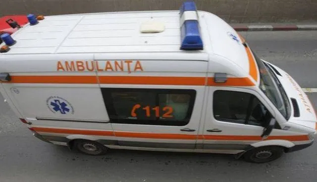 O ambulanță albă cu dungi portocalii și girofaruri albastre este parcată pe un drum asfaltat.