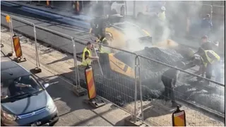 Mai mulți muncitori cu veste reflectorizante lucrează pe o suprafață de asfalt proaspăt întinsă, lângă un utilaj galben și un gard metalic.