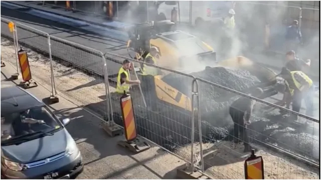Mai mulți muncitori cu veste reflectorizante lucrează pe o suprafață de asfalt proaspăt întinsă, lângă un utilaj galben și un gard metalic.
