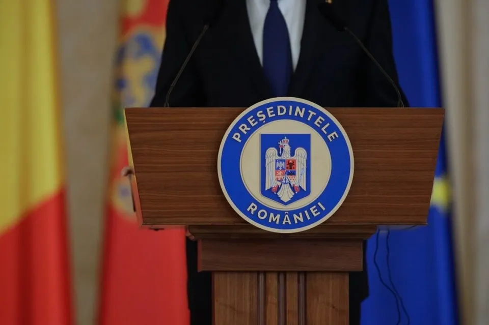 Un bărbat într-un costum închis și cravată albastră stă în spatele unui podium de lemn pe care este afișat emblema Președintelui României.