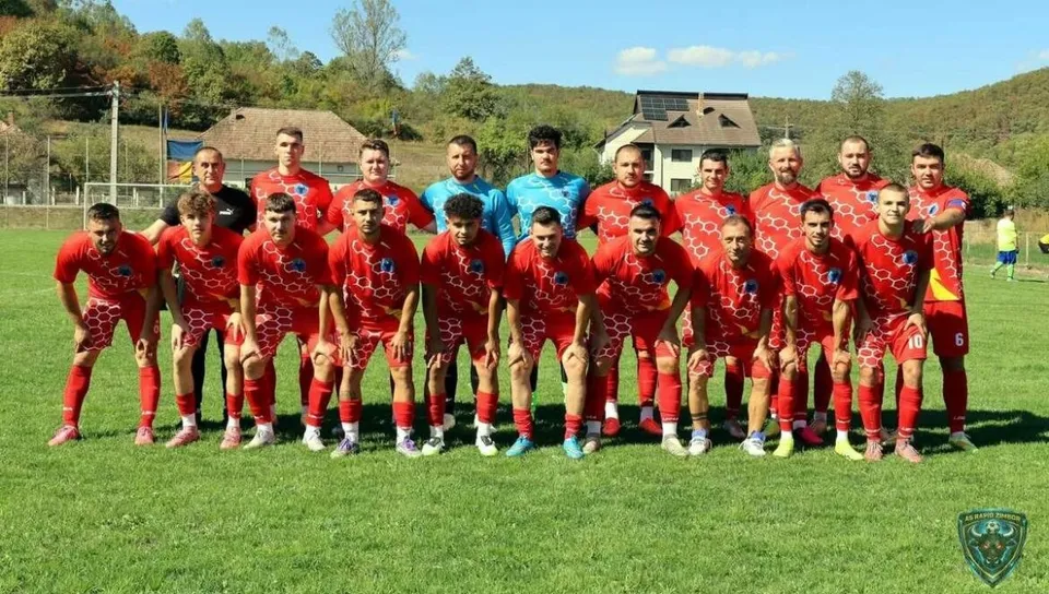 O echipă de fotbaliști în echipamente roșii posedă pentru o fotografie de grup pe un teren de fotbal verde sub un cer senin.