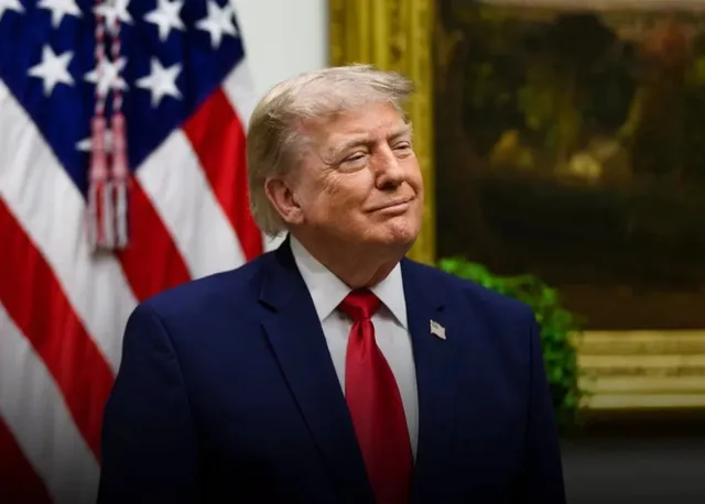 Un bărbat cu păr blond, purtând un costum albastru, cămașă albă și cravată roșie, stă în fața unui steag american.