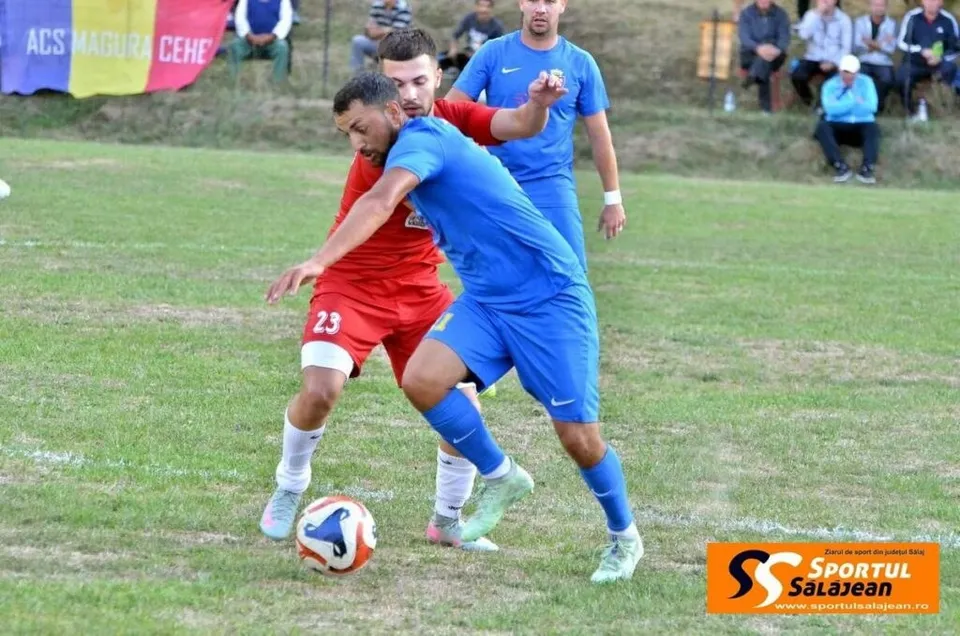 Trei jucători de fotbal concurează pe un teren de iarbă, unul purtând echipament roșu și celălalt echipament albastru, lângă un balon.