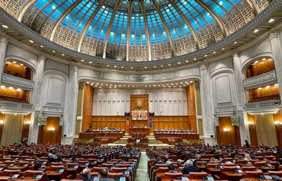 Interiorul unei săli circulare cu un plafon de sticlă decorat, containe de tribune din lemn și rânduri de scaune ocupate de persoane.