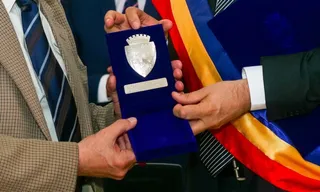 Două persoane țin o cutie de catifea albastră deschisă care conține un medalion argintiu cu un blazon sculptat.