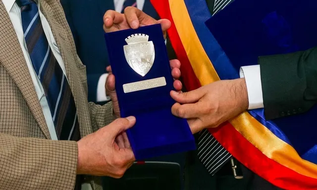 Două persoane țin o cutie de catifea albastră deschisă care conține un medalion argintiu cu un blazon sculptat.