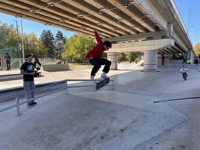 Un bărbat cu tricou roșu și pantaloni închiși execută un trick pe un skateboard deasupra unei bare metalice într-un skatepark de beton.