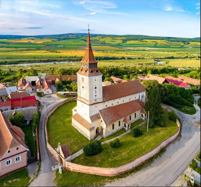 O biserică cu un turn înalt și acoperiș în formă de piramidă este înconjurată de un zid de incintă pe un teren cu iarbă verde.