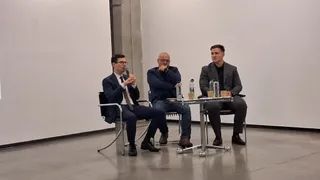 Trei bărbați stau așezați pe scaune lângă o masă mică într-o încăpere cu pereți albi.