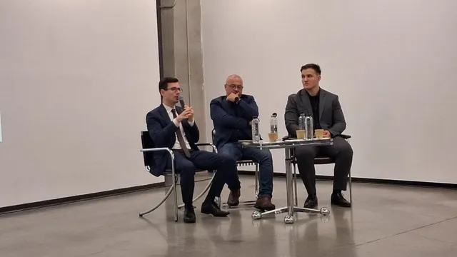 Trei bărbați stau așezați pe scaune lângă o masă mică într-o încăpere cu pereți albi.