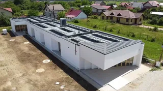 O clădire albă cu acoperiș înclinat și panouri solare este situată într-o zonă rurală cu case și vegetație în fundal.