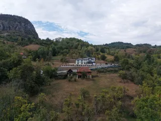 O pensiune albă cu terasă și balustradă stă pe un deal împădurit, lângă o construcție mai veche și o stâncă abruptă.