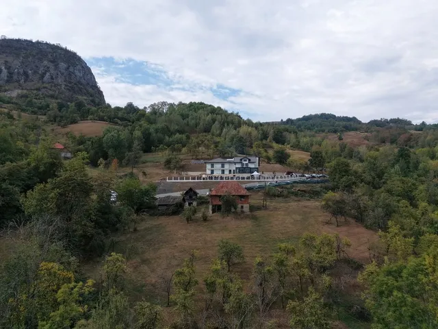O pensiune albă cu terasă și balustradă stă pe un deal împădurit, lângă o construcție mai veche și o stâncă abruptă.