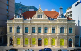 O clădire veche cu fațadă galbenă, acoperiș de țiglă roșie și ferestre cu rame albastre este situată sub un deal verde sub un cer senin.