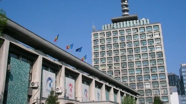 O clădire înaltă cu multe ferestre este situată lângă o construcție mai joasă care prezintă panouri verticale cu logo-uri colorate.