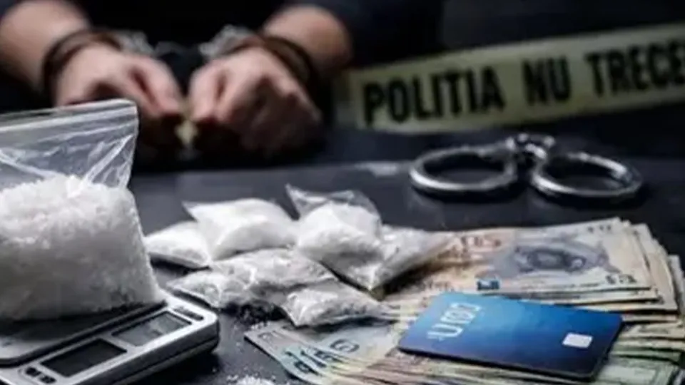 Mănuși de piele, cătușe, saci cu o substanță albă, bani și un card stau pe o suprafață întunecată lângă o bandă de poliție.