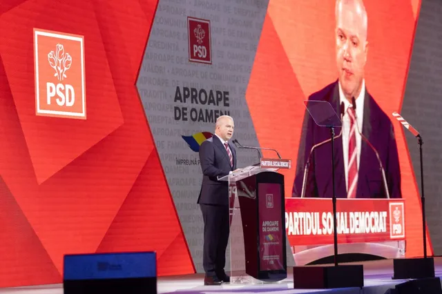 Un bărbat într-un costum închis la culoare stă în spatele unui podium și vorbește într-un microfon în fața unui ecran mare.