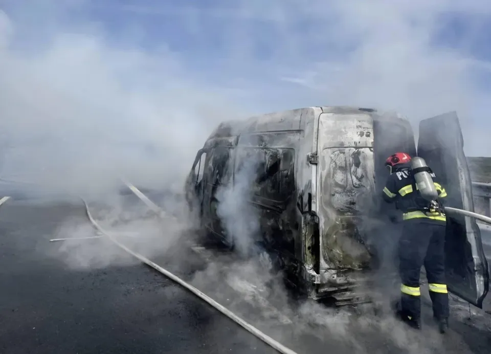 Un pompier cu echipament de protecție și cască roșie stă lângă o autoutilitară carbonizată din care se ridică fum pe un drum asfaltat.