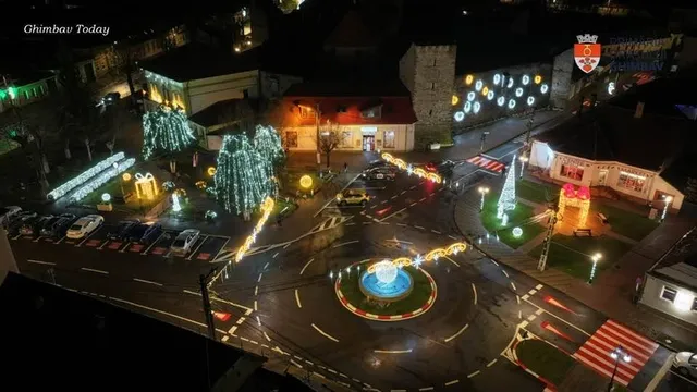 Aerial night view of a town square decorated with illuminated Christmas trees, light displays, and a central roundabout fountain.