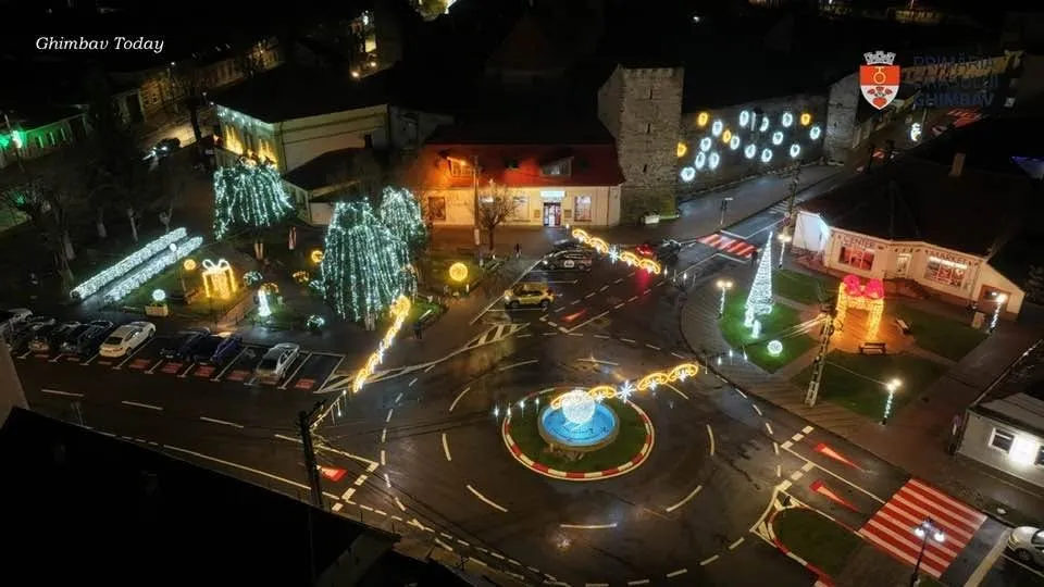 Aerial night view of a town square decorated with illuminated Christmas trees, light displays, and a central roundabout fountain.
