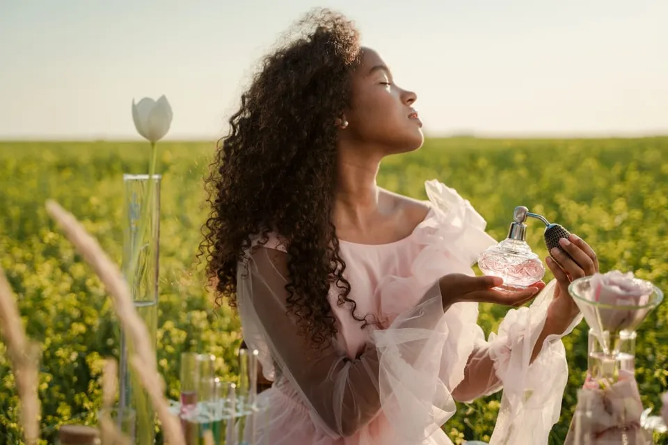 O femeie cu părul crepusculat, purtând o rochie roz, ține un flacon de parfum într-o câmpie verde sub lumină naturală.