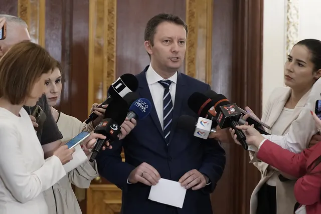 Un bărbat într-un costum albastru și cravată strie la microfoane ținute de mai multe persoane în fața unui fundal din lemn.