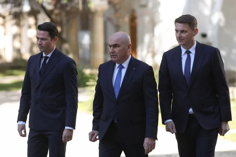 Trei bărbați în costume închise și cravate merg pe o cărare lângă o clădire cu coloane.