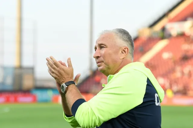 Un bărbat cu părul scurt și grij, purtând un tricou de culoare verde deschis și negru, aplaudă pe un teren de fotbal.