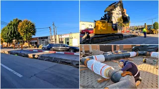 Un excavator galben lucrează pe un șantier lângă o mașină neagră, în timp ce niște țevi mari sunt așezate pe pământ lângă un om.