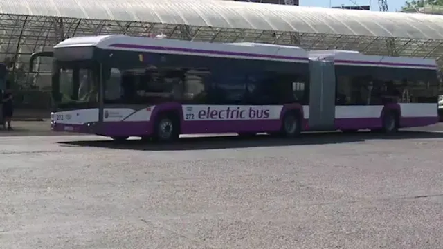Un autobuz articulat alb cu dungi violete și textul electric bus este parcat pe un teren asfaltat sub o structură acoperită cu material alb.