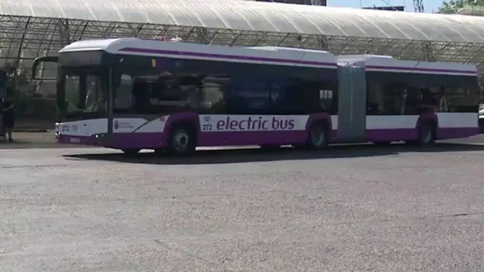Un autobuz articulat alb cu dungi violete și textul electric bus este parcat pe un teren asfaltat sub o structură acoperită cu material alb.