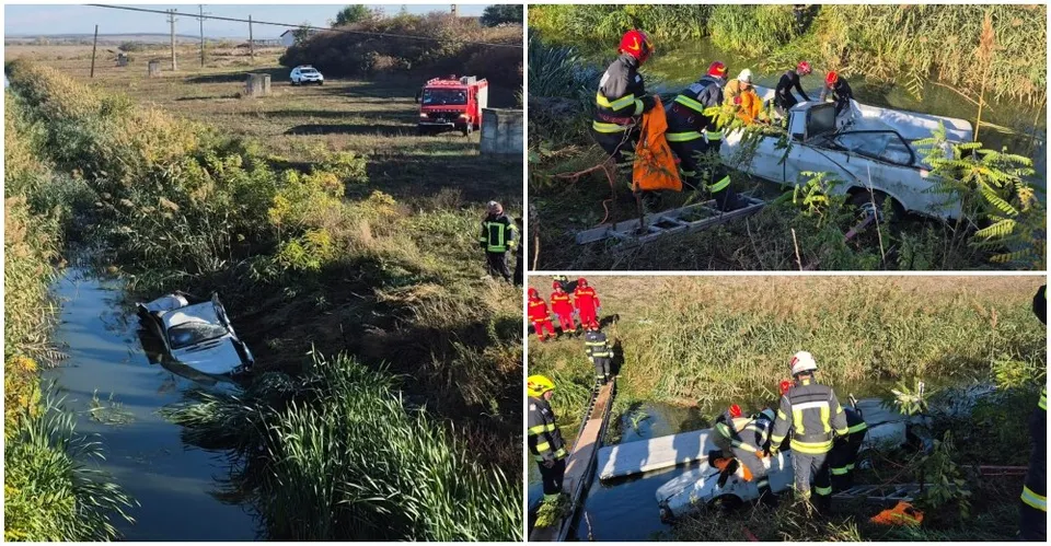 Pompieri în echipamente reflectorizante lucrează lângă un autoturism alb parțial scufundat într-un canal îngust înconjurat de vegetație.