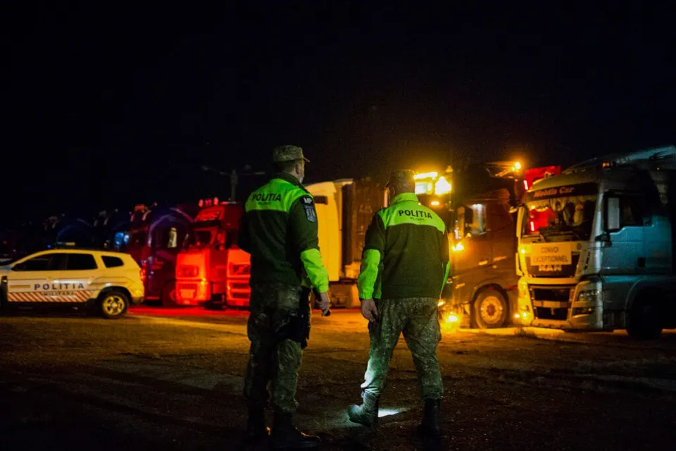 Două persoane în uniforme verzi și camuflaj stau pe un drum noaptea, lângă mașini de poliție și camioane parcate.